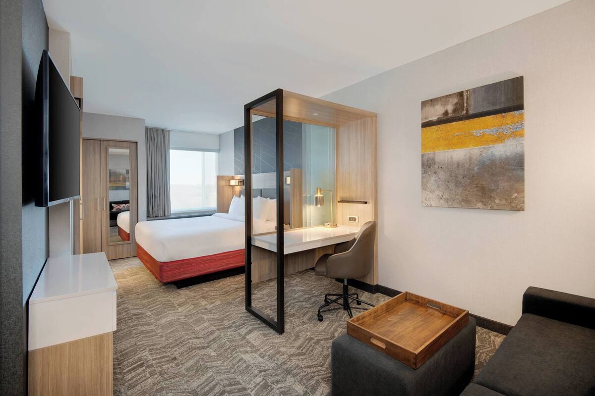 A hotel room with a bed, a desk, a chair, a TV, and a painting on the wall.