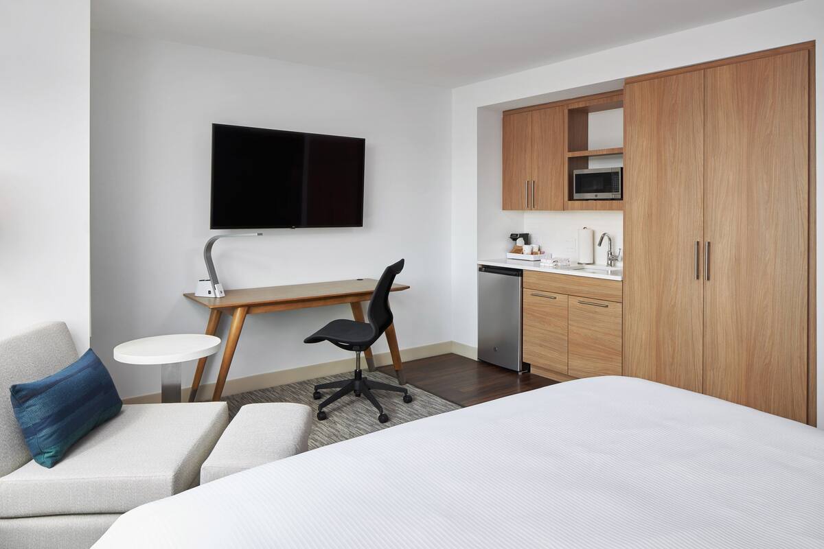 Camera d'albergo con un letto, una scrivania con una sedia, una televisione, un forno a microonde e armadietti in legno.