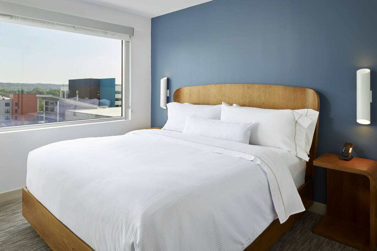 Una moderna camera d'hotel con un ampio letto, una testata imbottita in legno e vista sulla città attraverso una grande finestra.