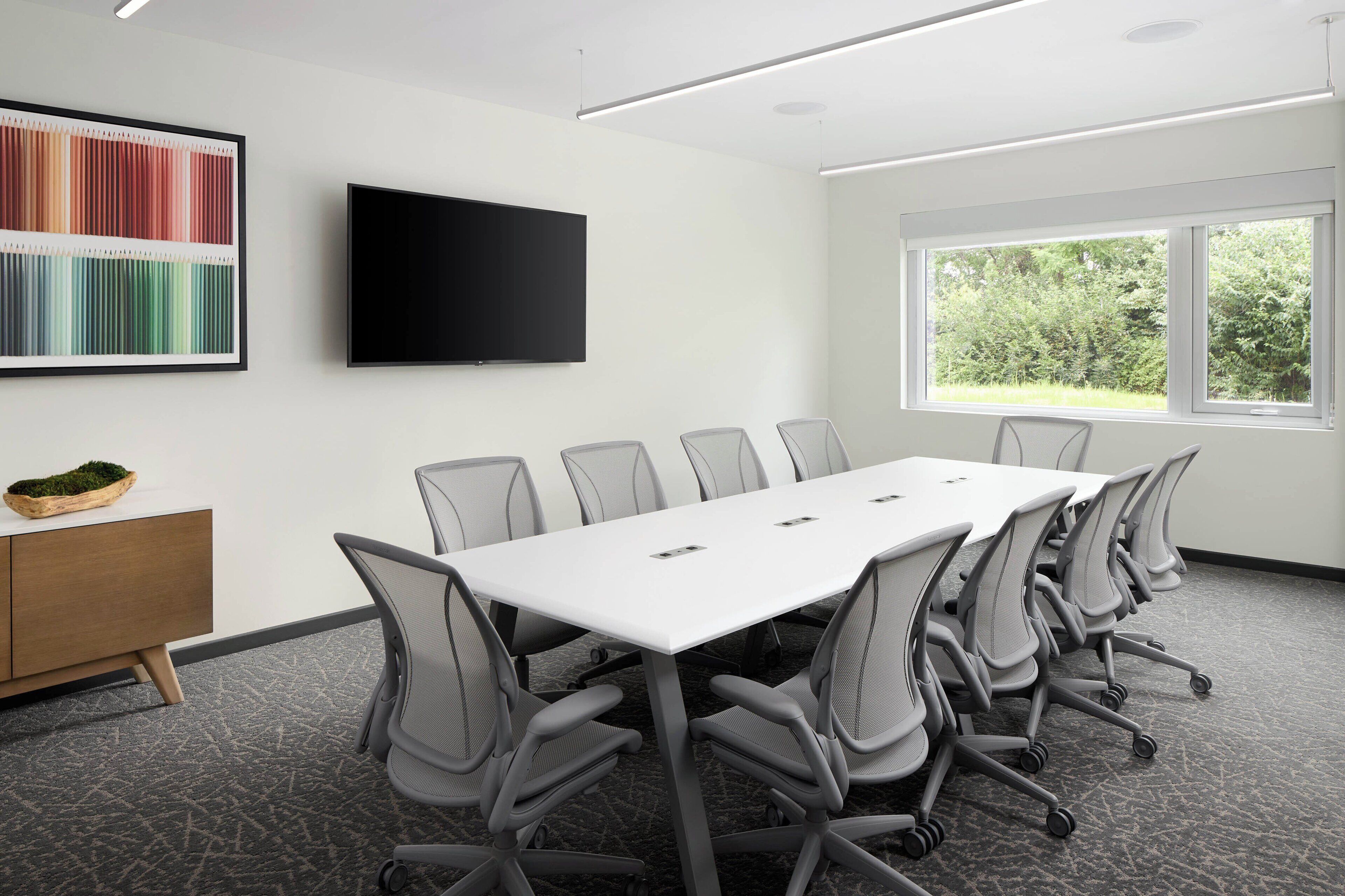 Una sala de conferencias con una mesa larga, sillas, un televisor de pantalla plana, un ventanal con vista a árboles y un elemento decorativo en la pared.