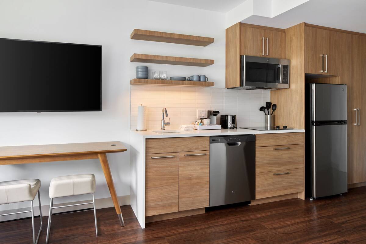 Una cucina moderna con mobili in legno, un frigorifero in acciaio inossidabile, un forno a microonde e una lavastoviglie. C'è un tavolo da pranzo in legno con due sedie e un televisore a schermo piatto montato a parete.