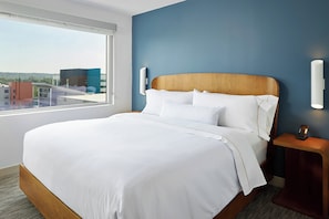 A hotel room with a large bed, a nightstand, a wall-mounted lamp, and a window with a city view.