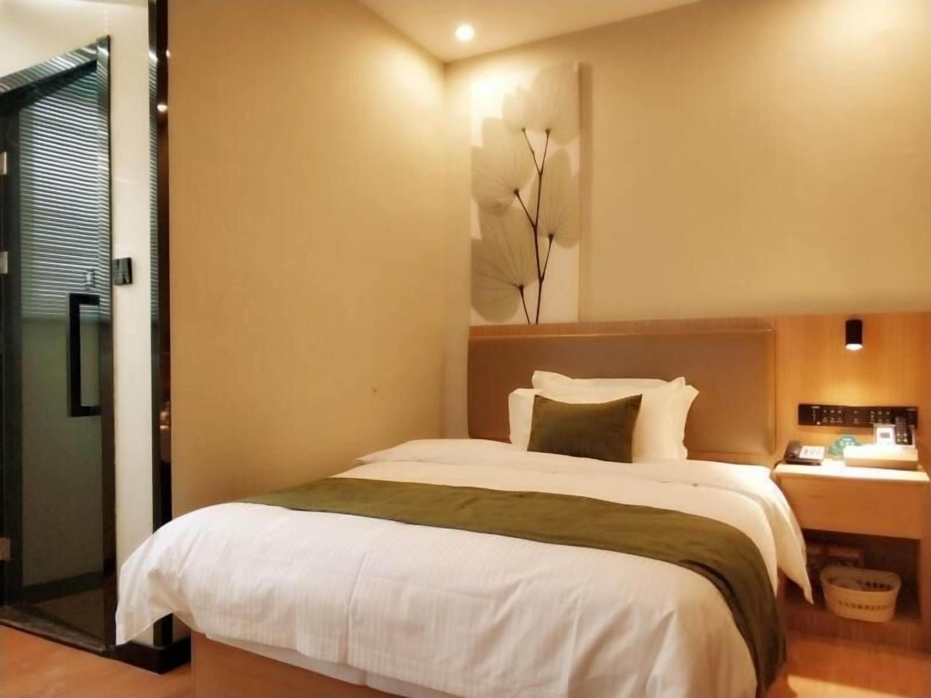 A hotel room with a bed, a nightstand, a wall-mounted lamp, and a door with blinds.