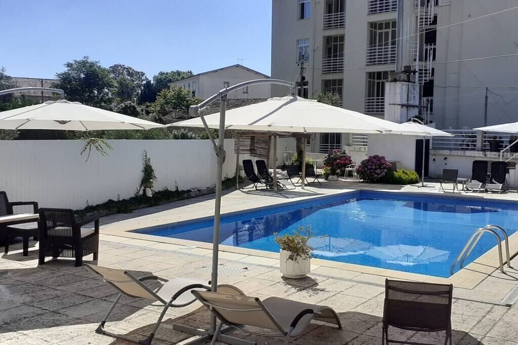 A swimming pool with lounge chairs and umbrellas, surrounded by a white wall and a building in the background.