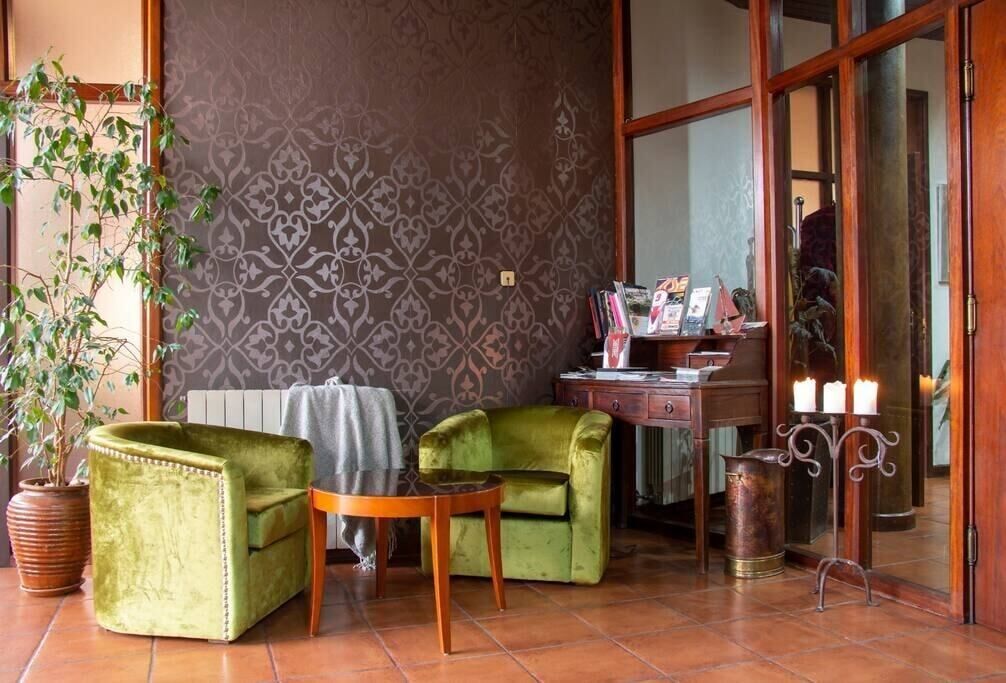 Una habitación acogedora con dos sillones verdes, una pequeña mesa de madera, un radiador, un escritorio de madera con revistas, una planta en maceta y un candelabro.