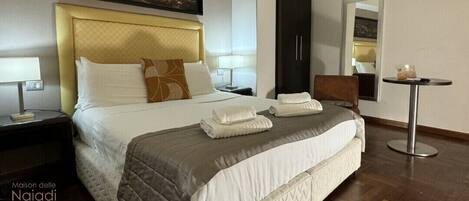 A neatly made bed with a headboard, a bedside table with a lamp, a small round table with a glass, and a framed picture on the wall.