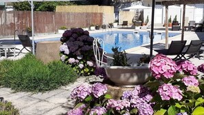Piscina con sillones de descanso y sombrillas, rodeada de plantas en macetas y flores.