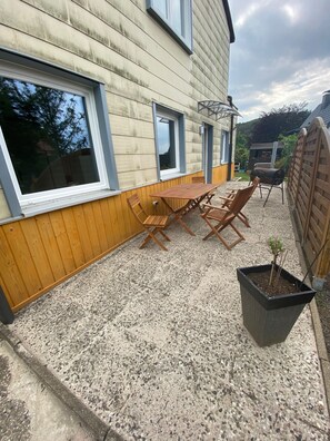 Eine Terrasse mit einem Holztisch, Stühlen, einem kleinen Topfpflanzen und einem Grill.