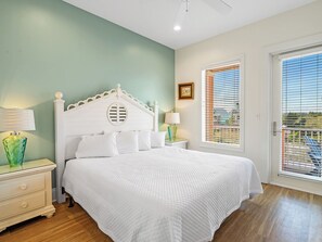 A bedroom with a white bed, a nightstand with a lamp, a window with blinds, and a door.