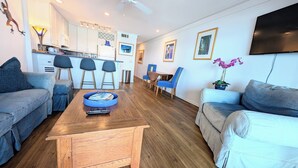 A modern living room with a wooden coffee table, two sofas, a kitchen in the background, and a TV mounted on the wall. - Seamist #6-Oceanfront-Wonderful View! (Seacrest)