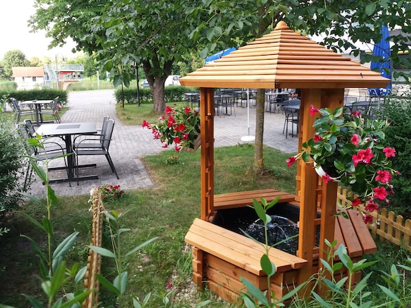 A wooden gazebo with a bench, surrounded by greenery and a small pond.