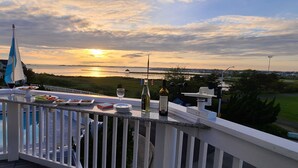 Un balcón con vista al atardecer sobre el agua, una bandera y una mesa con platos y vasos.