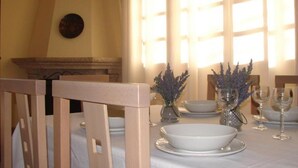Een eethoek met een tafel voor vier personen, voorzien van witte borden, kommen en wijnglazen. Op tafel staan twee vazen met lavendel, en op de achtergrond een stenen openhaard.
