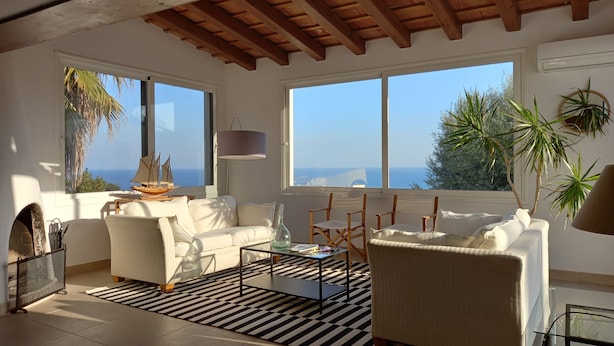 A spacious living room with a high ceiling, large windows offering ocean views, a fireplace, and a variety of seating options.