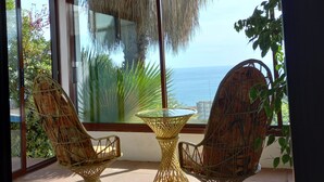 A covered patio with wicker furniture and a glass table, overlooking a scenic view of the ocean.