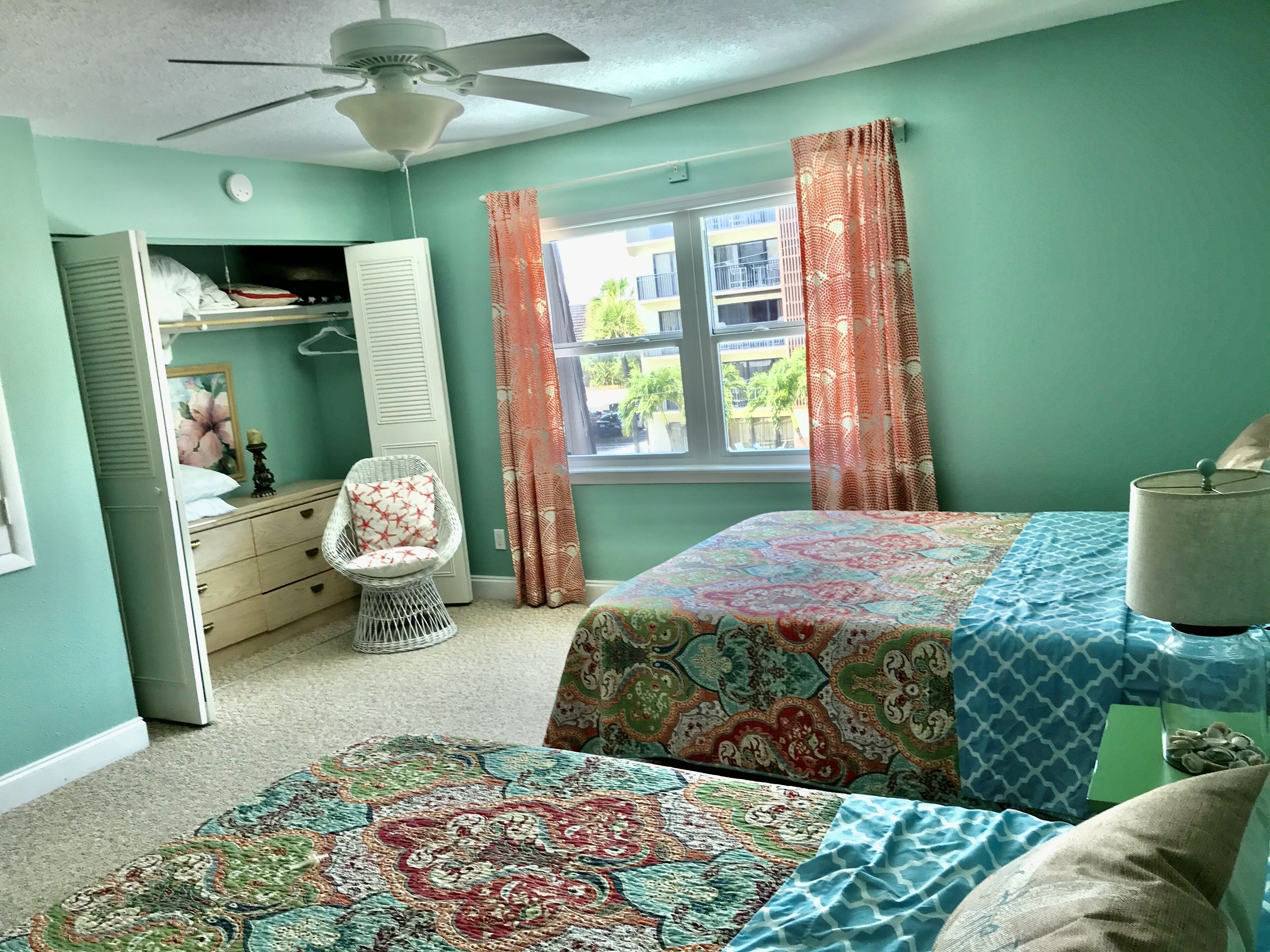 Une chambre avec deux lits, une chaise, un ventilateur de plafond, une fenêtre avec des rideaux et un radiateur.