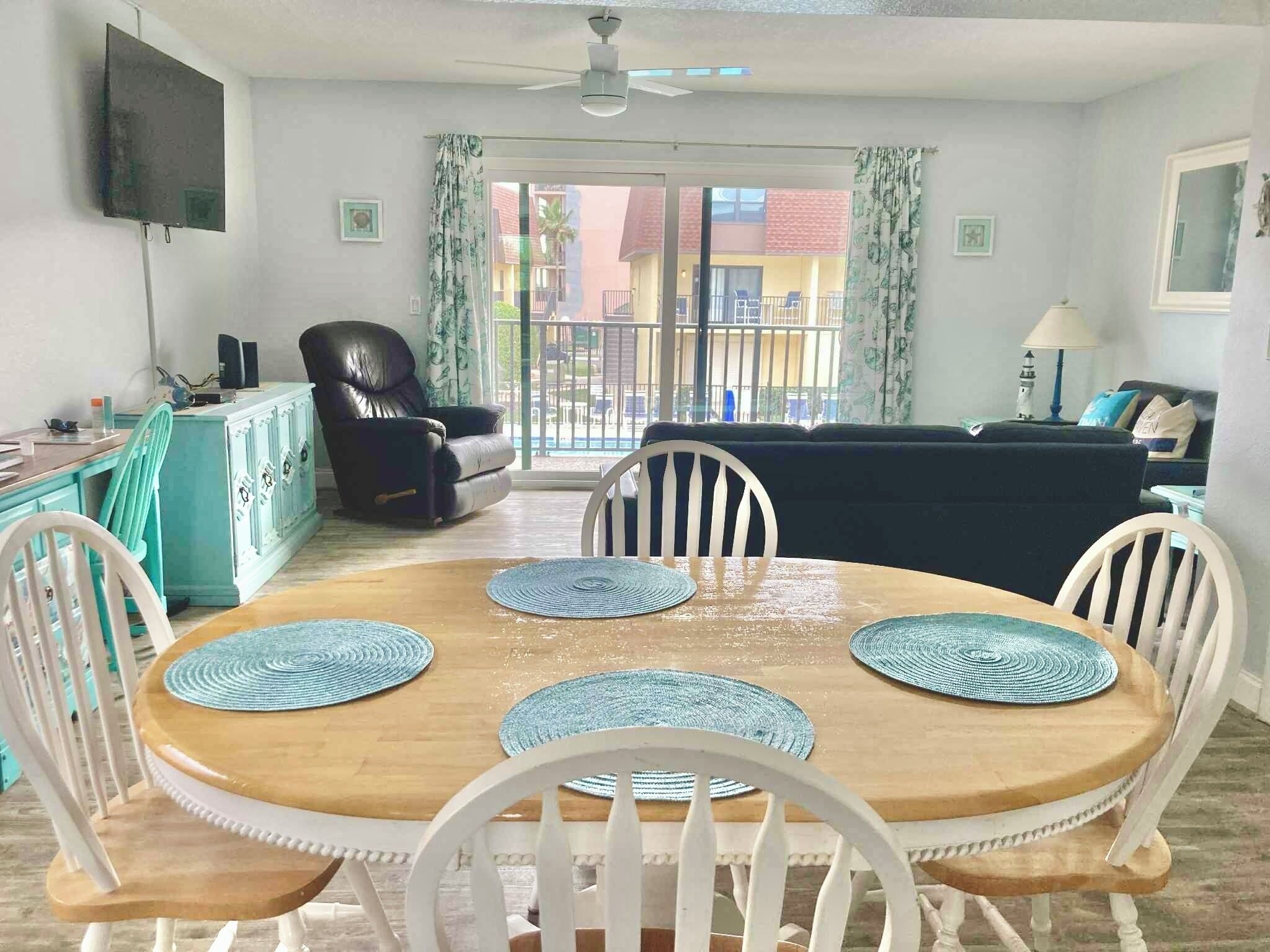 A dining area with a wooden table and white chairs, a black leather chair, a teal cabinet, and a TV mounted on the wall.