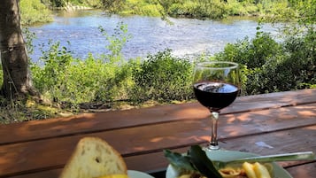 A picnic table with a bowl of fruit and pasta, a glass of red wine, and a view of a river and forest.