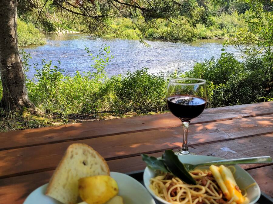 A picnic table with a bowl of fruit and pasta, a glass of red wine, and a view of a river and forest.