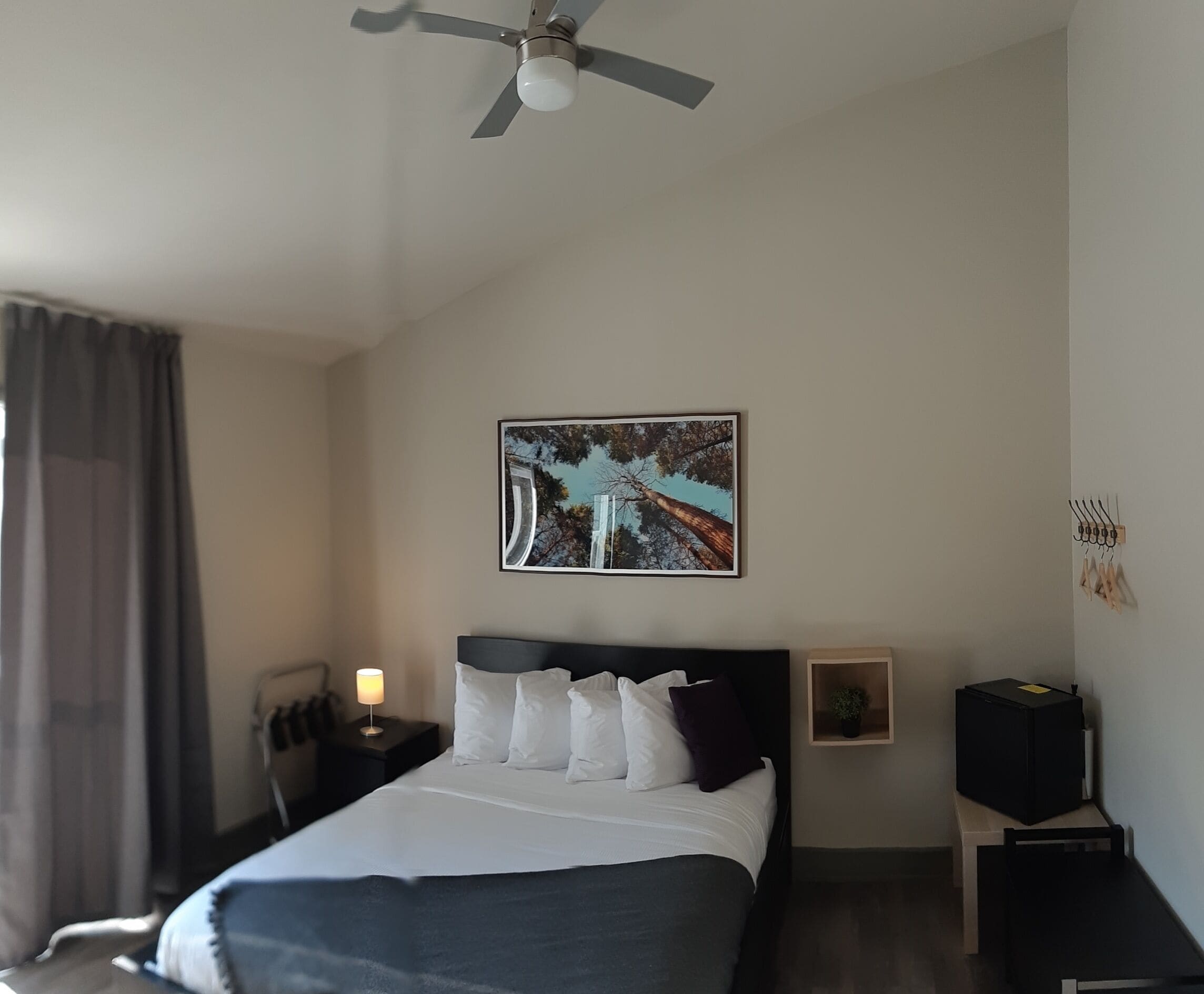 Una camera da letto con un letto, una ventola a soffitto, un comodino con una lampada, una TV a muro e un quadro incorniciato.