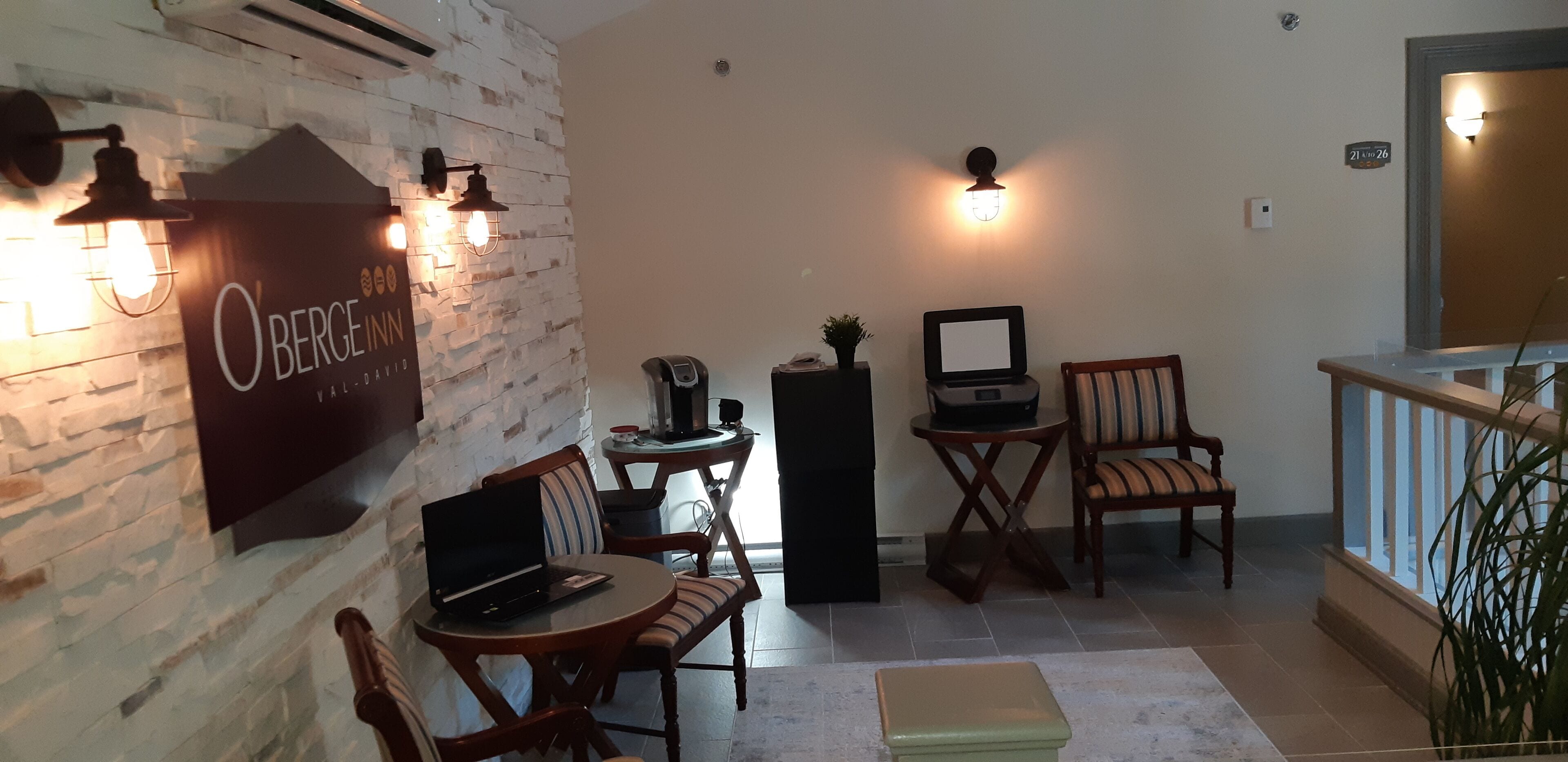 Een gezellige kamer met een stenen muur, een laptop op een tafel, twee stoelen, een kleine tafel met een telefoon en een plant, en een bordje met de tekst 'OBERGE INN'.
