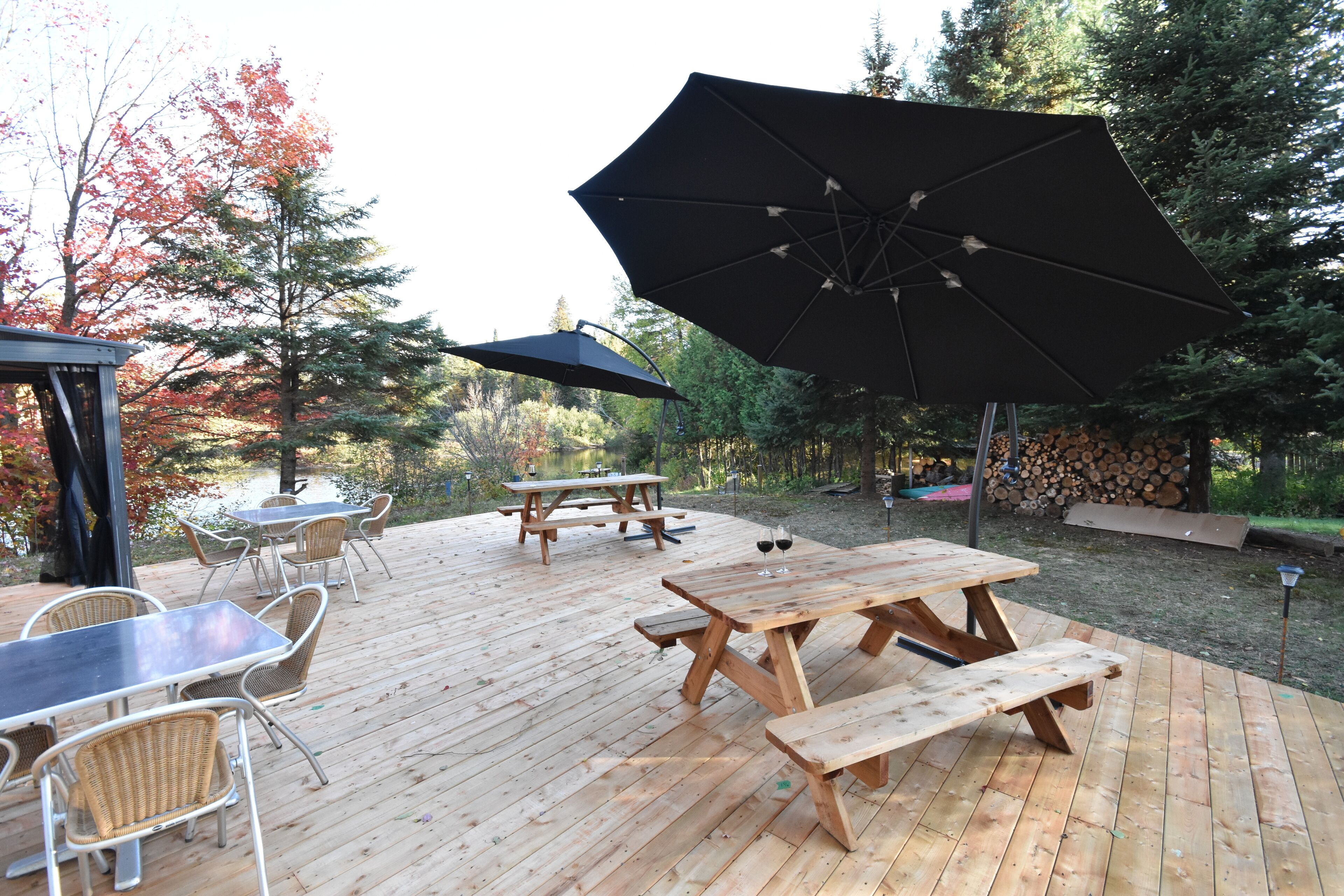 A wooden deck with picnic tables and chairs, surrounded by trees and a stack of firewood.
