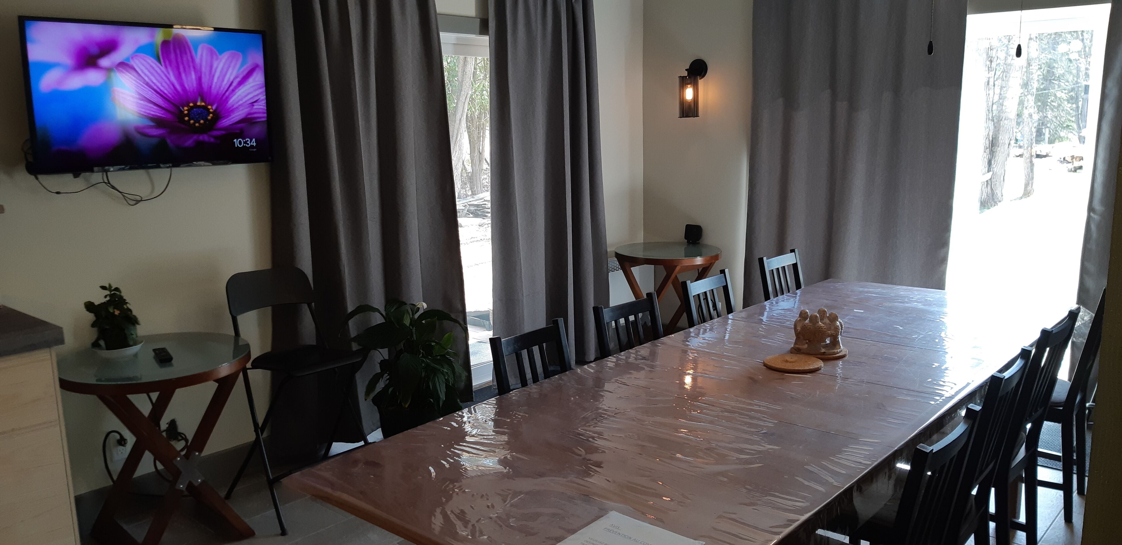 A dining area with a long table, chairs, a small round table, and a television on the wall.