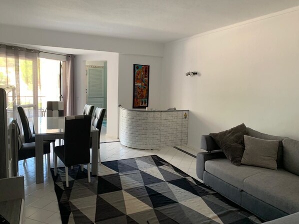 A modern living room with a grey sofa, a dining area with a white table and chairs, and a kitchen area with a white brick backsplash.