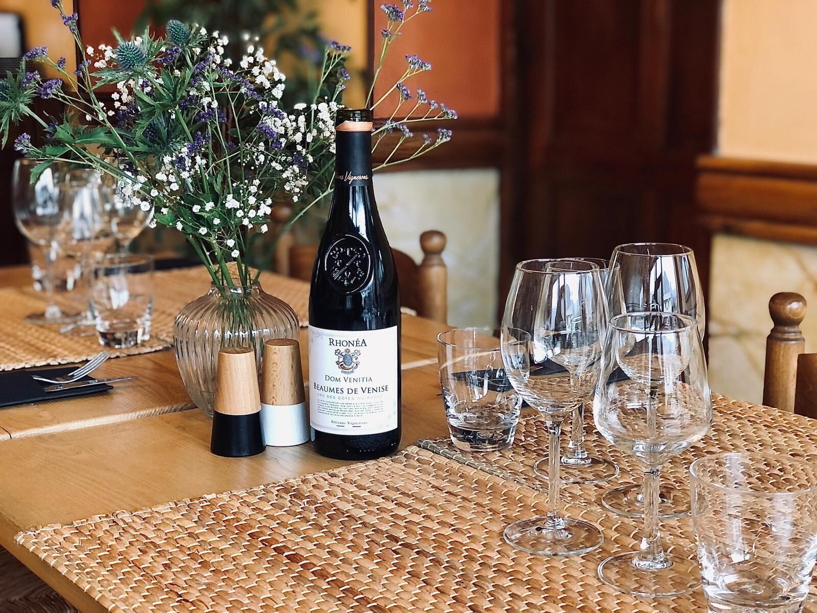A table set with wine glasses, a bottle of RhoÃ©na Dom Venitia Beaumes de Venise, and a vase with flowers.