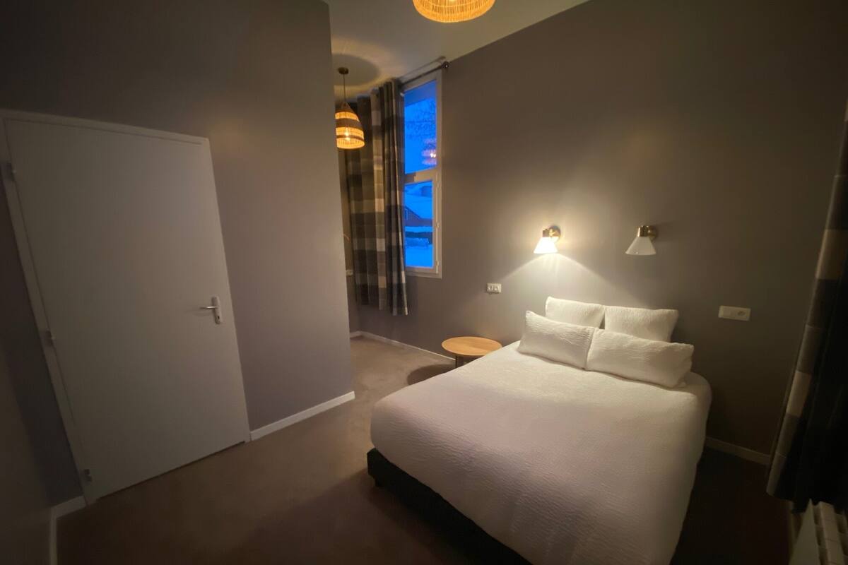 A bedroom with a large bed, a bedside table, a wall-mounted lamp, and a window with curtains.