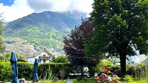 A garden with outdoor seating, a large tree, and a mountain in the background. - Hotel Oberland (Le Bourg-d'Oisans)
