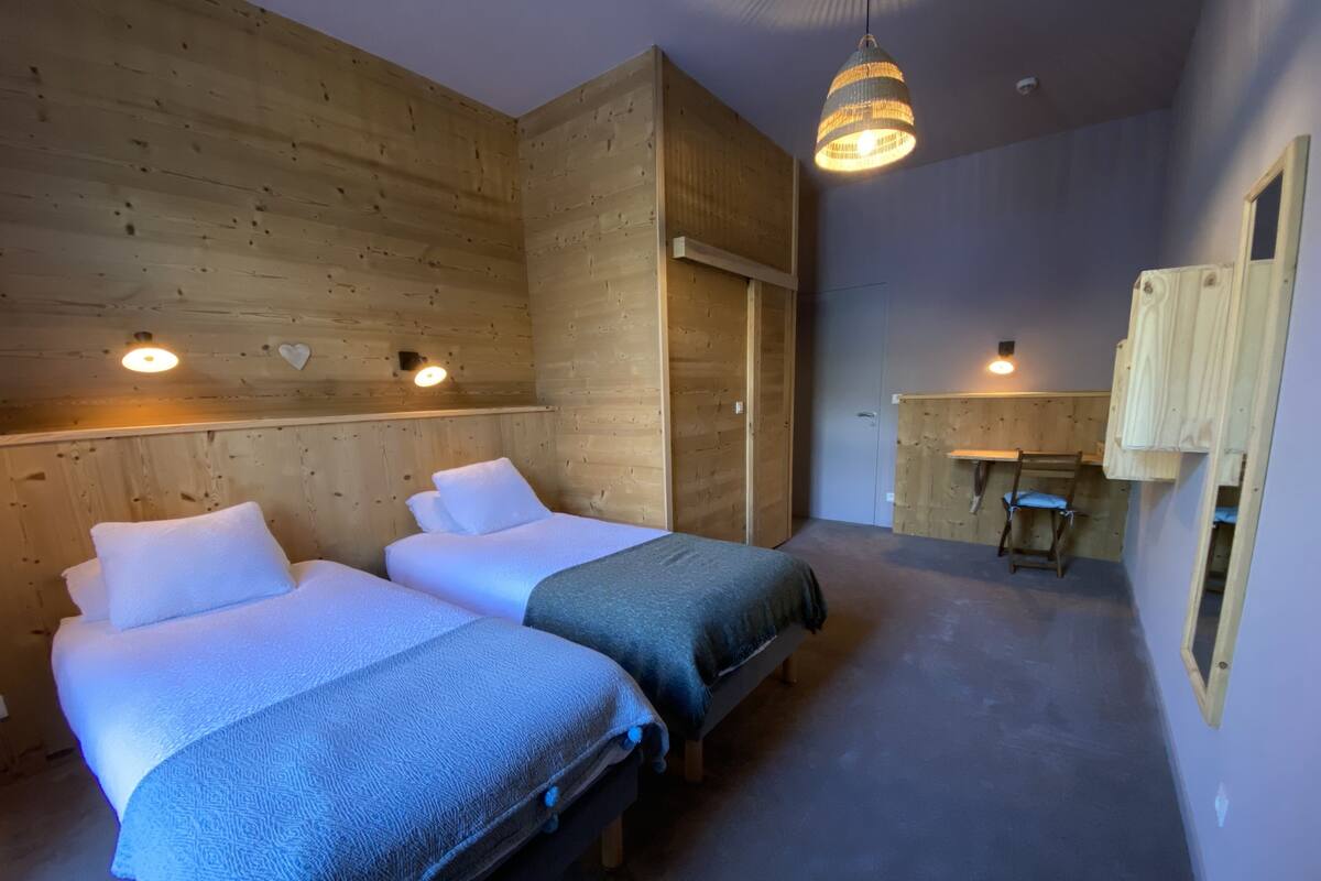 A room with two beds, wooden walls, a small desk, and a hanging light fixture.