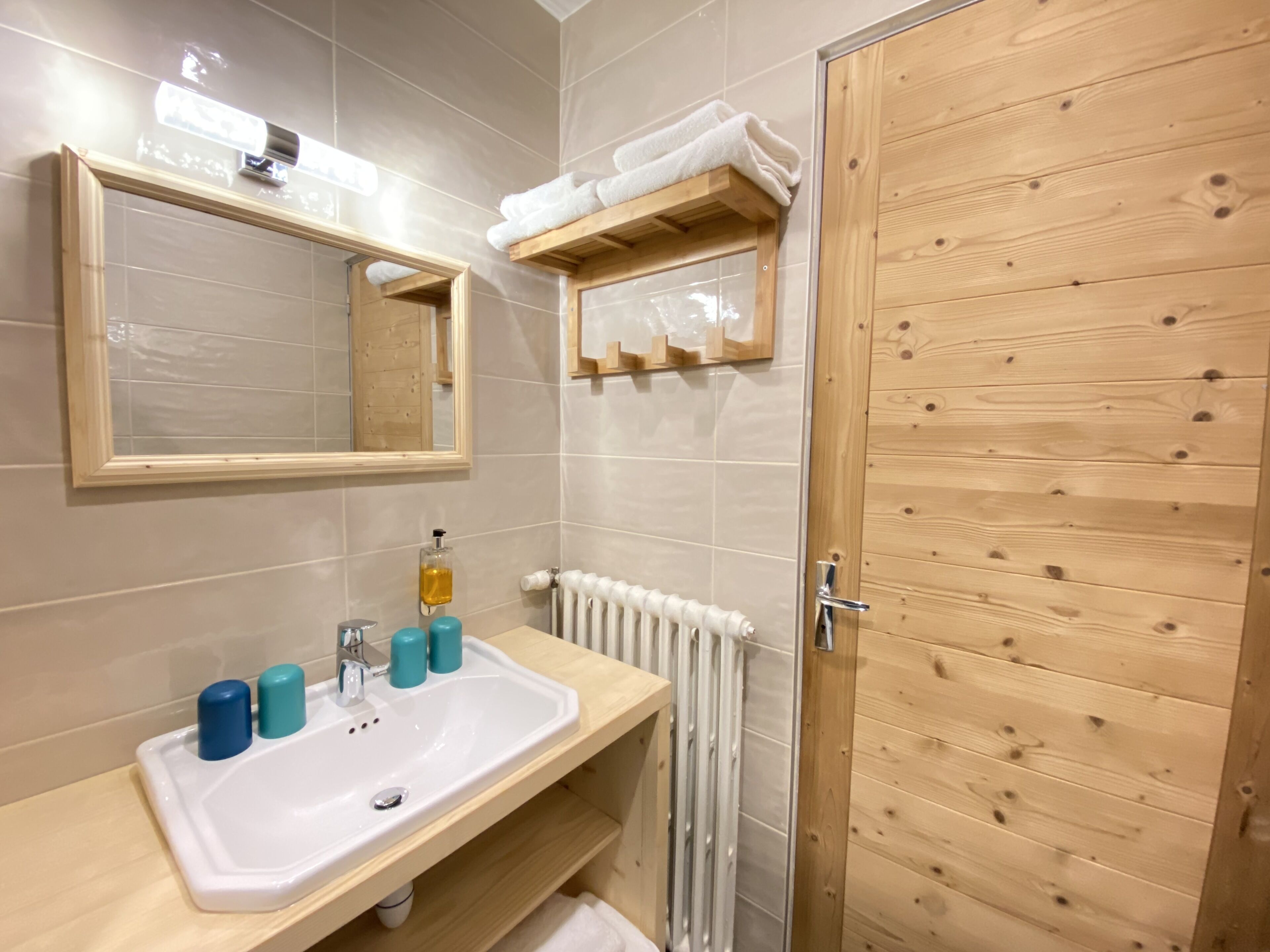 Un baño con puerta de madera, un lavamanos blanco con dispensadores de jabón azules, un espejo y un perchero para toallas.