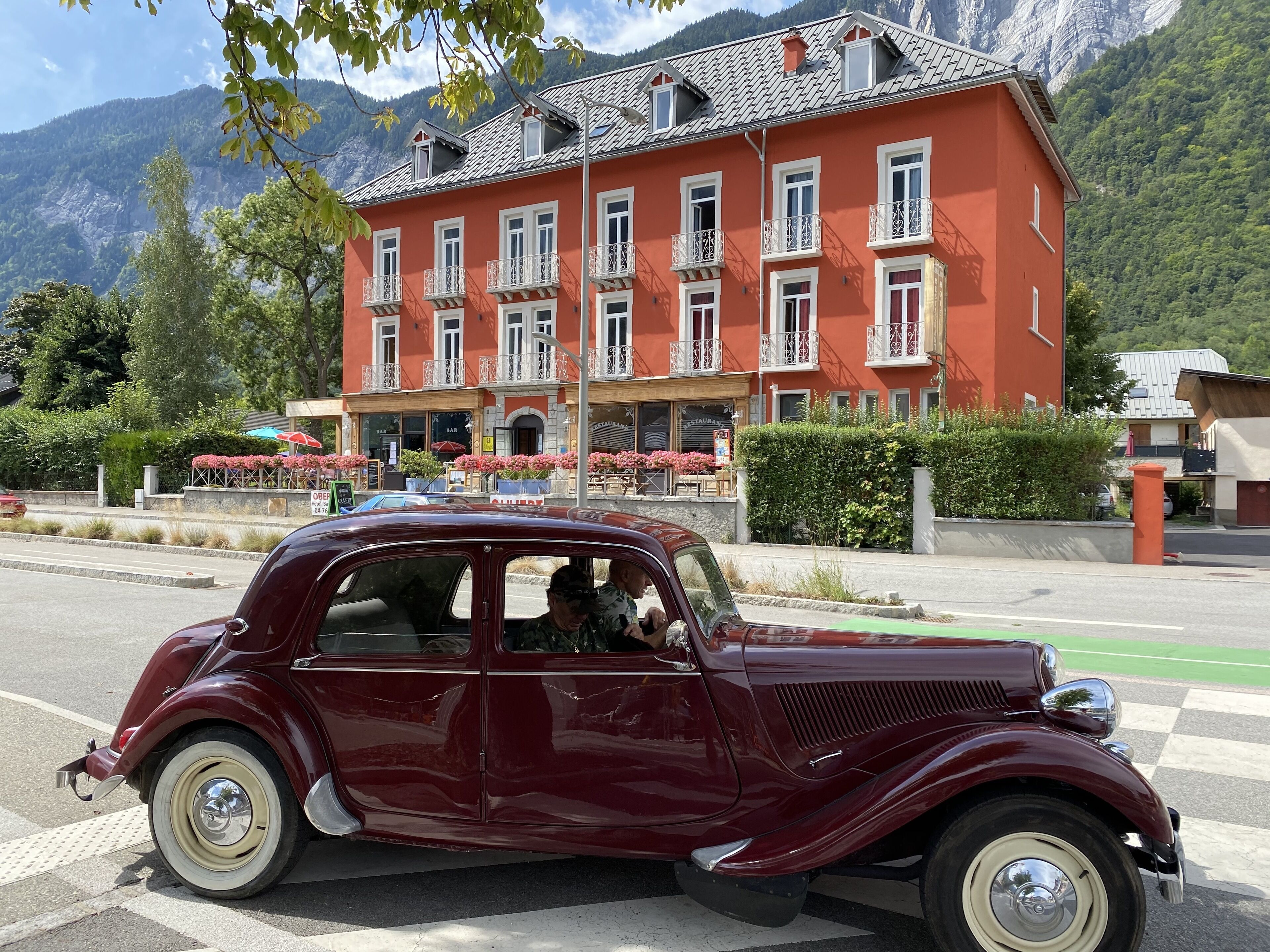 A vintage red car parked in front of a red building with balconies and a mountain backdrop.