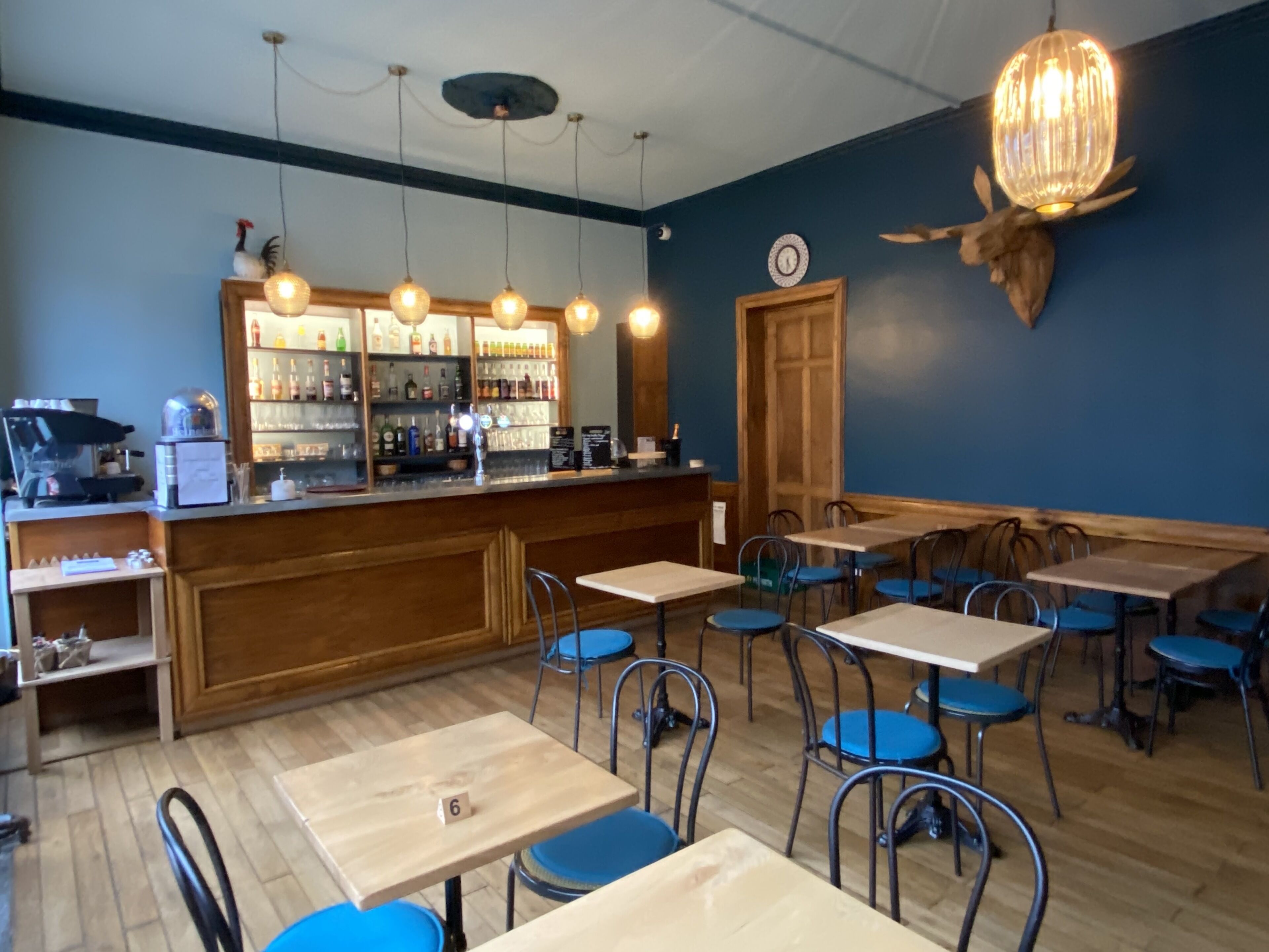 A restaurant interior with wooden tables and chairs, a bar area with shelves of bottles, and a decorative wall piece.