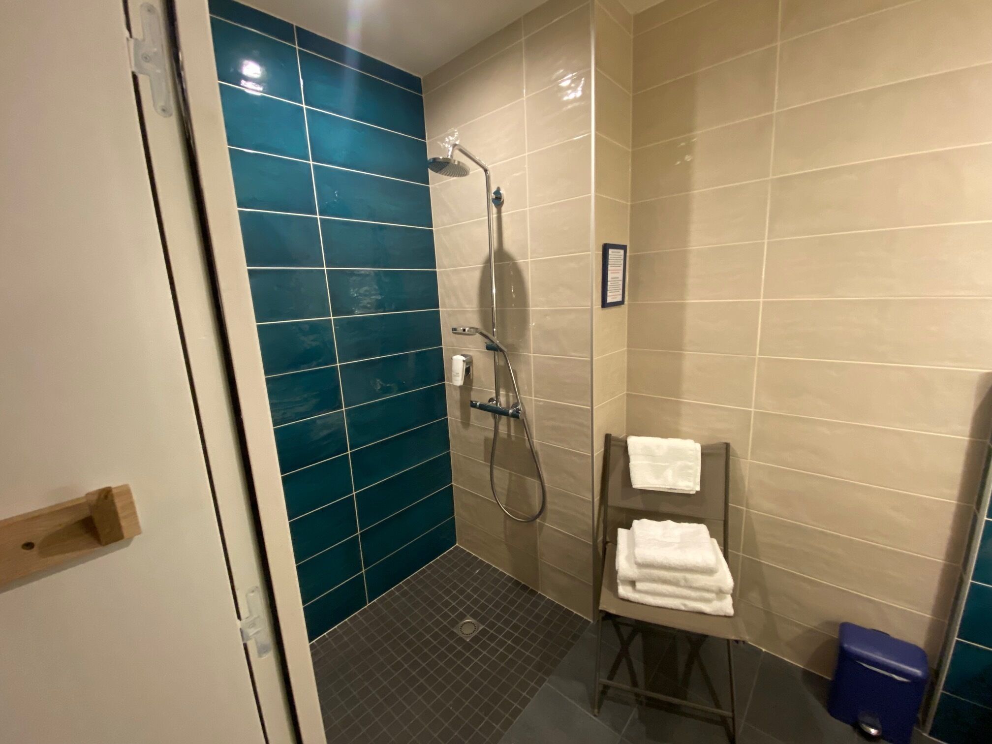 A small, tiled shower area with a handheld showerhead, a towel rack with folded towels, and a blue trash bin.