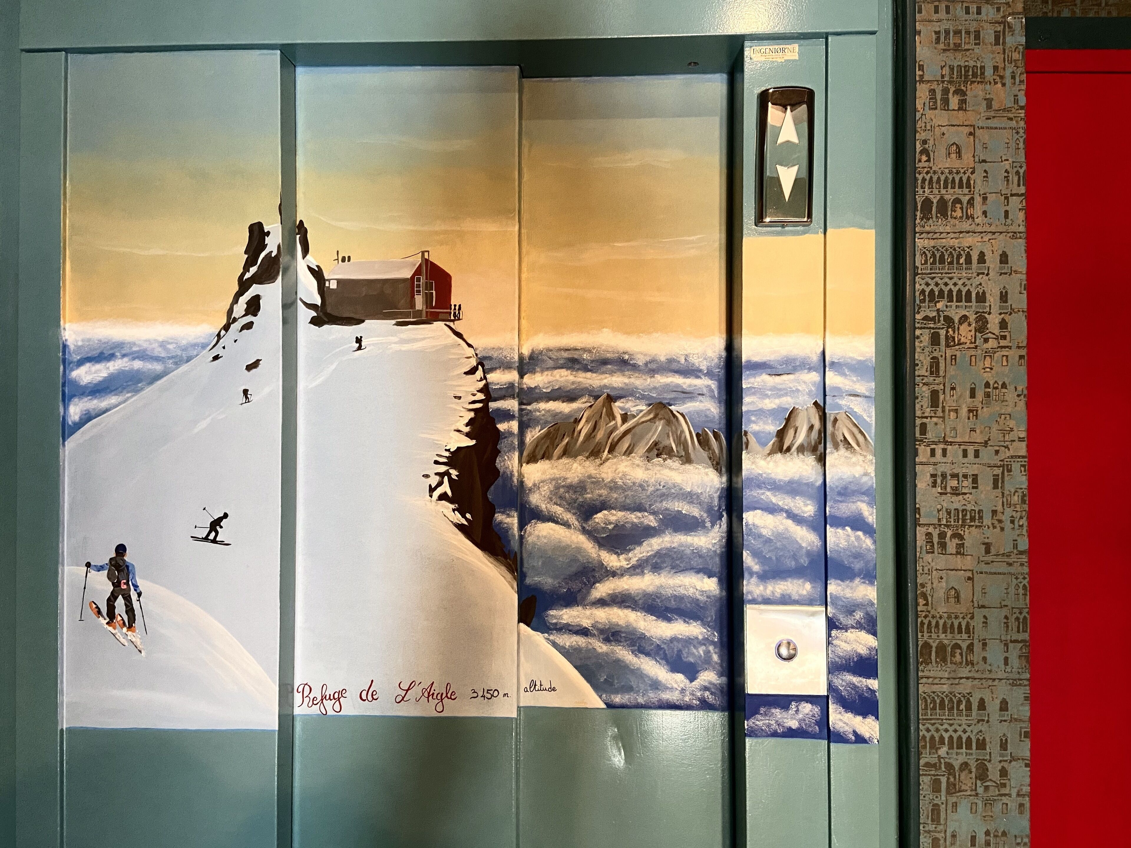 An elevator interior with a painting of a snowy mountain scene, including skiers and a red cabin.