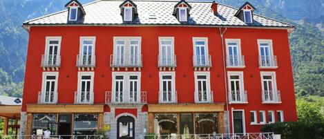 A red building with multiple windows and balconies, a central entrance, and a mountain backdrop.
