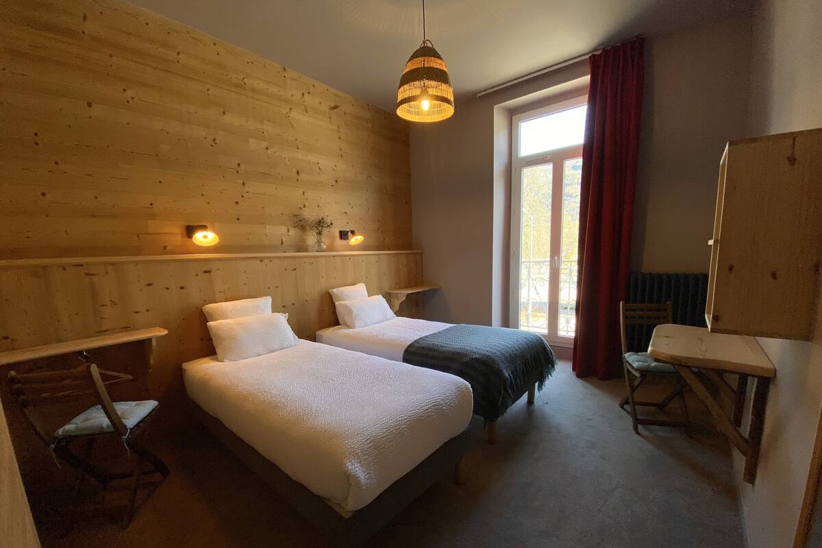 A room with two beds, a wooden wall, a hanging light fixture, a window with a view, and a chair.