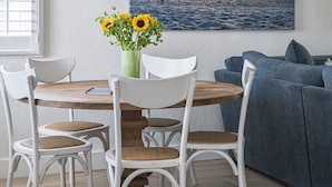 Un comedor con mesa de madera, sillas blancas, un jarrón con girasoles y un cuadro de atardecer colgado en la pared.