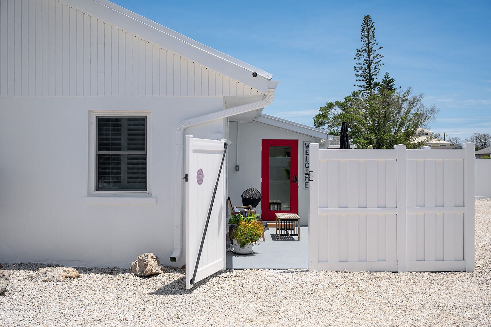 A white building with a red door, a window with shutters, and a gravel driveway.