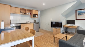 Une cuisine moderne avec des meubles en bois, un four à micro-ondes et une machine à café. On y trouve une table à manger avec des chaises et un canapé gris. Un téléviseur est posé sur un meuble en bois, et un tableau est accroché au mur.
