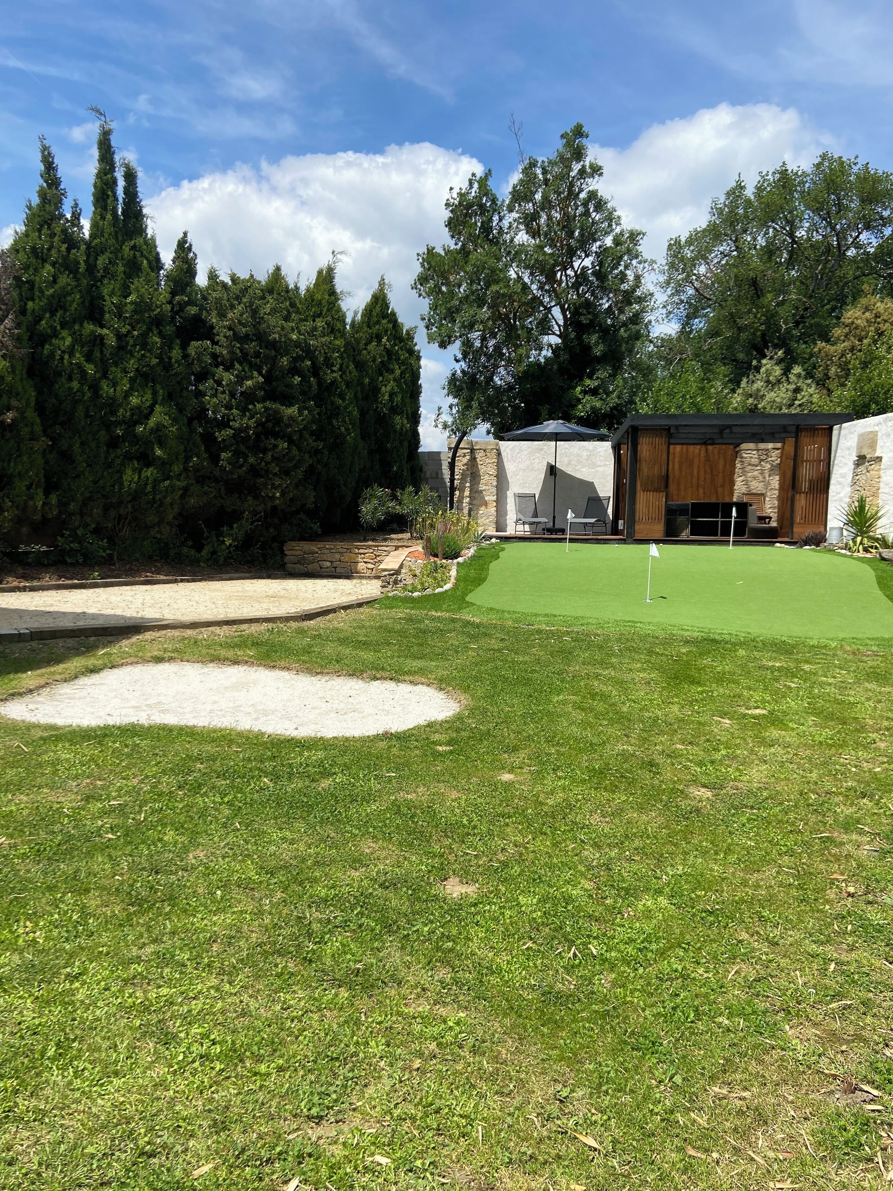 Un parcours de golf bien entretenu, doté d’un green d’entraînement, d’un bunker de sable et d’un espace de repos couvert avec des chaises et un parasol.