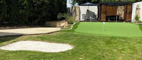 Un parcours de golf bien entretenu, doté d’un green d’entraînement, d’un bunker de sable et d’un espace de repos couvert avec des chaises et un parasol.