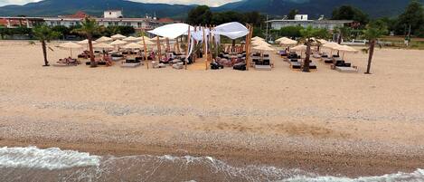 A beach with sun loungers and umbrellas, a pebble beach, and mountains in the background.