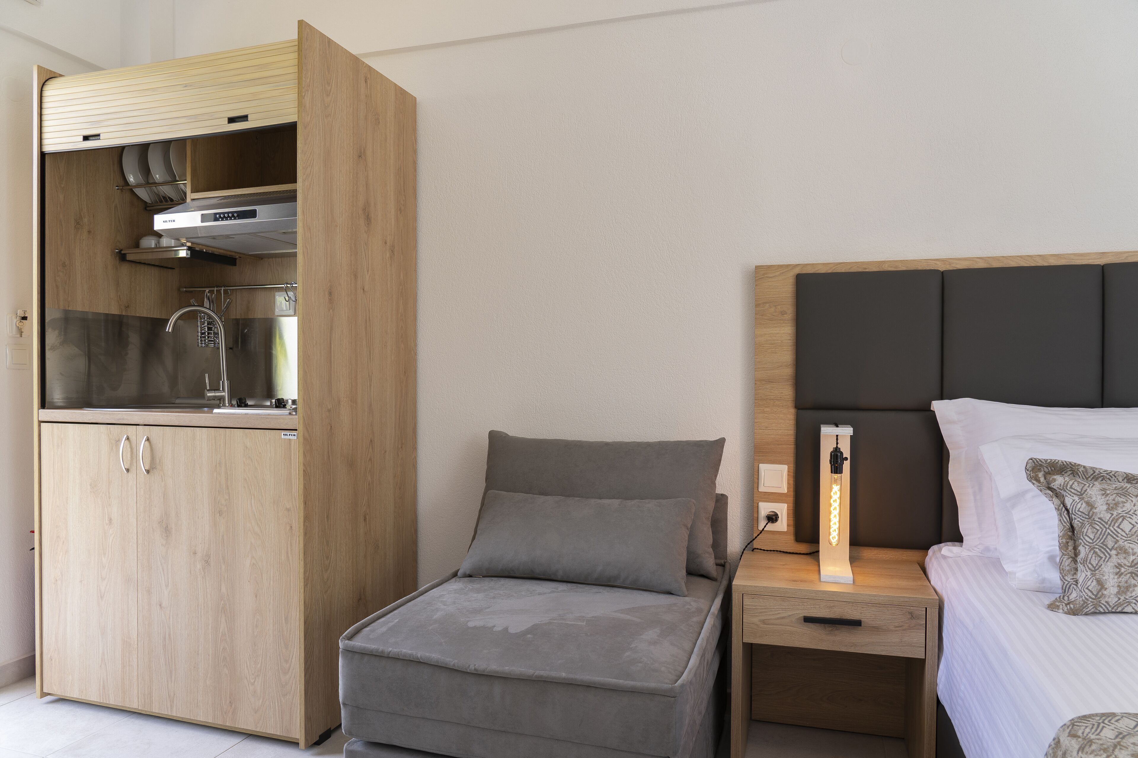 Une pièce compacte comprenant un coin cuisine, un lit avec une tête de lit et une table de chevet avec une lampe.