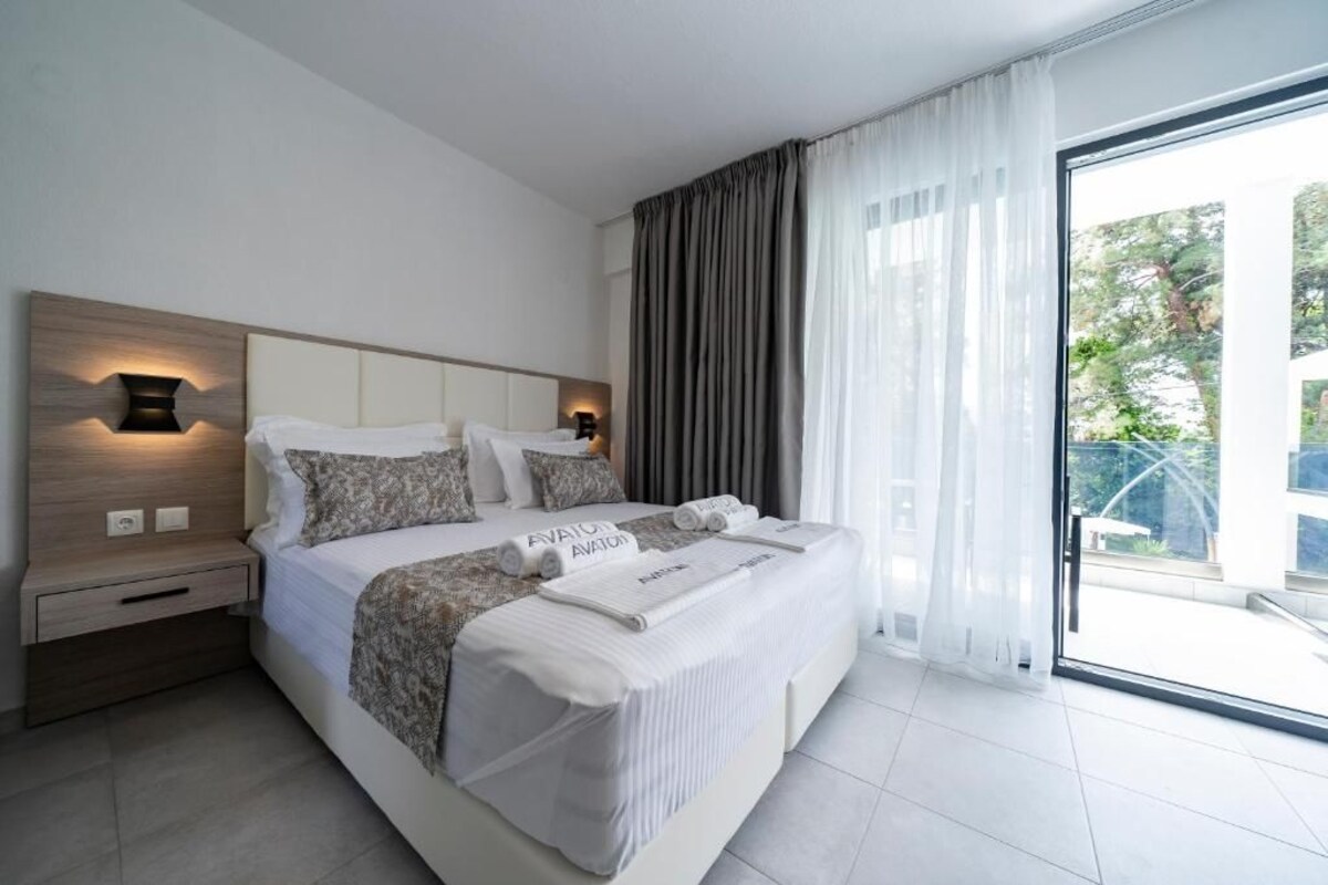 A modern hotel room with a large bed, a nightstand, a wall-mounted lamp, and a view through a glass door.