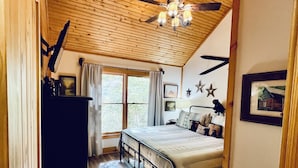 A bedroom with a wooden ceiling, a bed with a headboard, a desk, and a window with curtains. - Riverfront Cabin Downtown Helen! Hot Tub, FirePit,Arcade,Tubing & Private Park!♥️ (Helen)