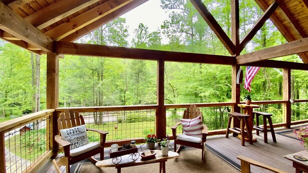 A wooden deck with a view of a forest. There are wooden chairs and tables, and an American flag is visible. - Riverfront Cabin Downtown Helen! Hot Tub, FirePit,Arcade,Tubing & Private Park!♥️ (Helen)