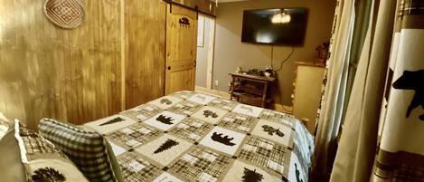Een slaapkamer met een houten wand, een plafondventilator, een gewatteerde dekbedovertrek, een televisie en een raam met gordijnen.